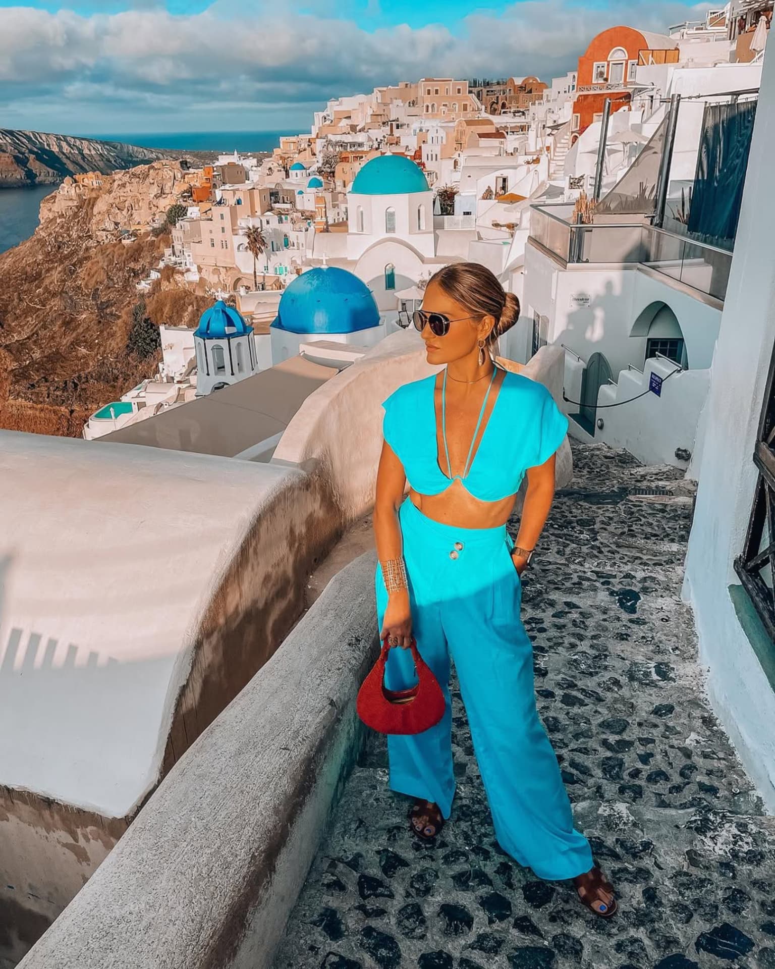 Emilim de conjunto turquesa numa escadaria de Santorini, com as cúpulas azuis ao fundo