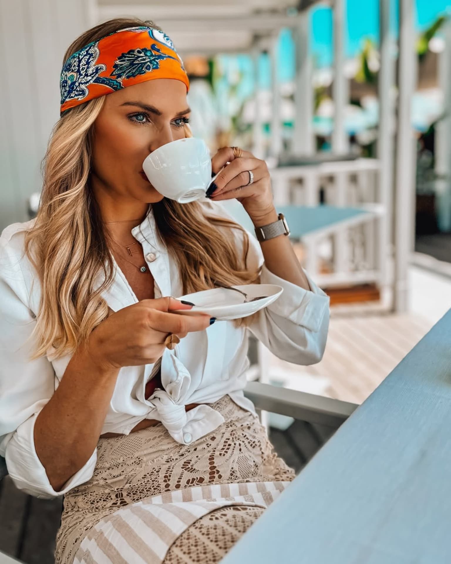 Emilim numa varanda de café à beira-mar, bebendo de uma xícara branca, com o mar turquesa ao fundo