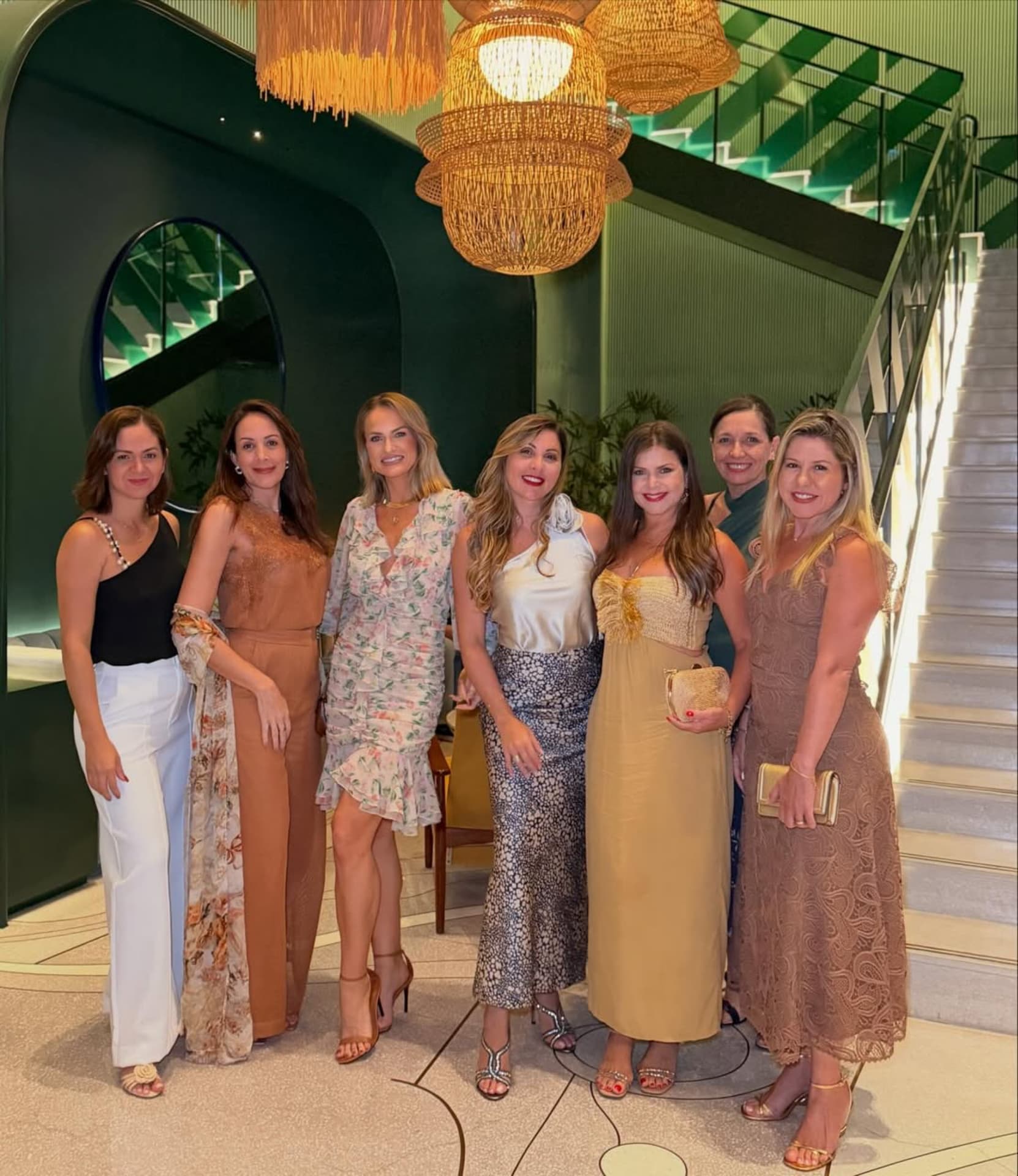 Emilim com o grupo de sete mulheres da expedição, em fila e de rosto, num saguão de paredes verdes com lustre de vime iluminado e escadaria ao fundo