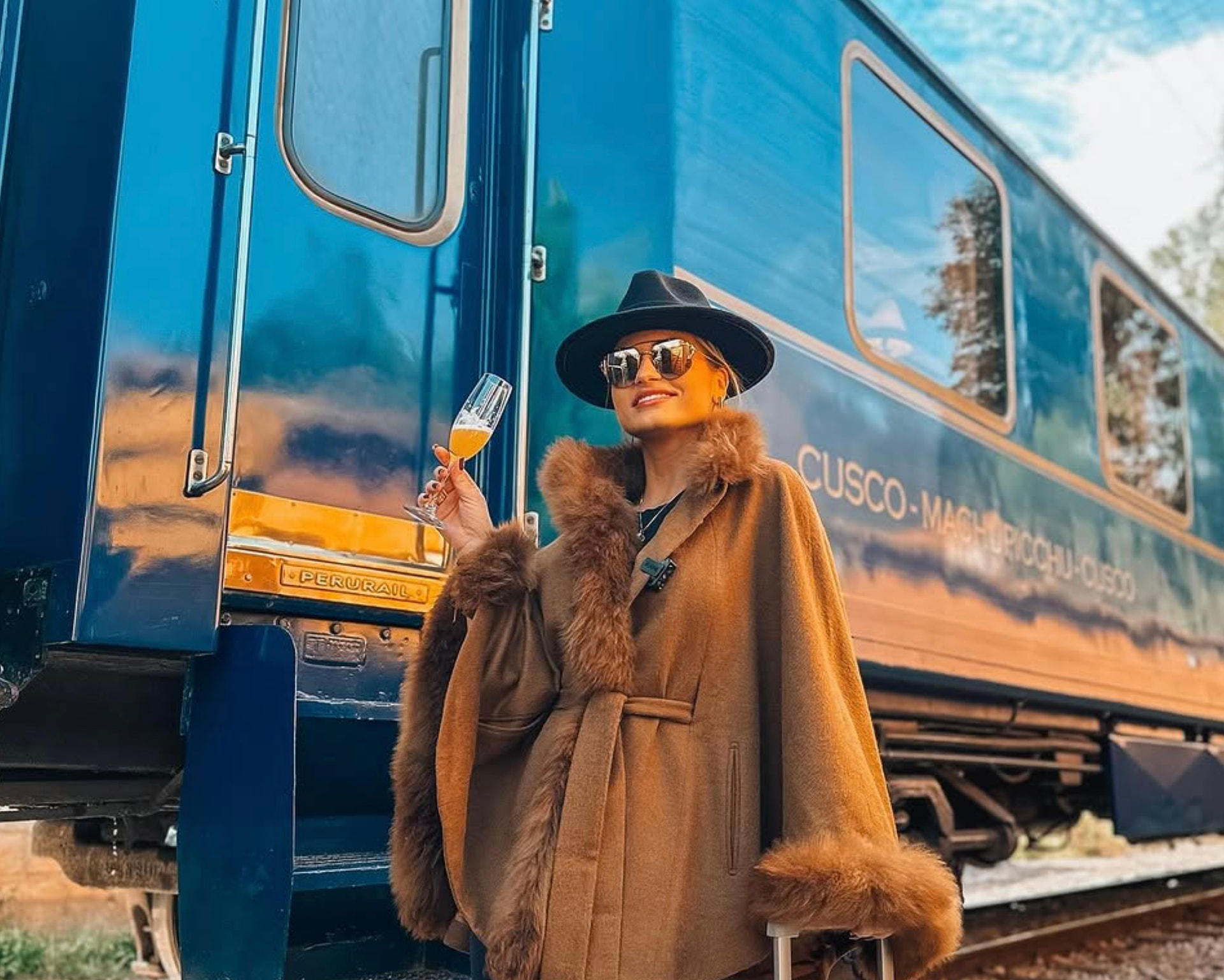 Emilim de chapéu preto e capa camel diante do trem da Perurail, com uma taça na mão e a mala ao lado, na rota de Cusco a Machu Picchu