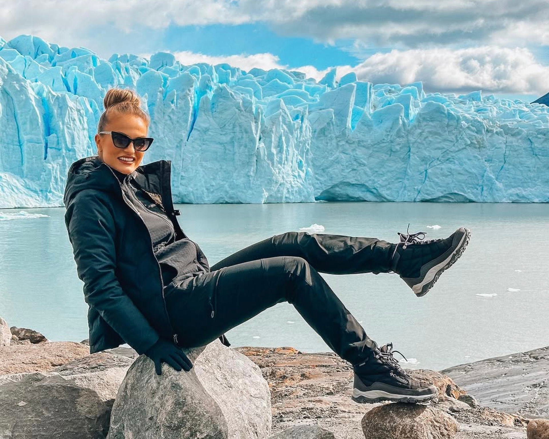 Emilim de jaqueta preta, óculos escuros e luvas, sentada nas pedras diante da parede azul de um glaciar, com uma bota de trilha apoiada numa rocha e a outra suspensa no ar, na Patagônia