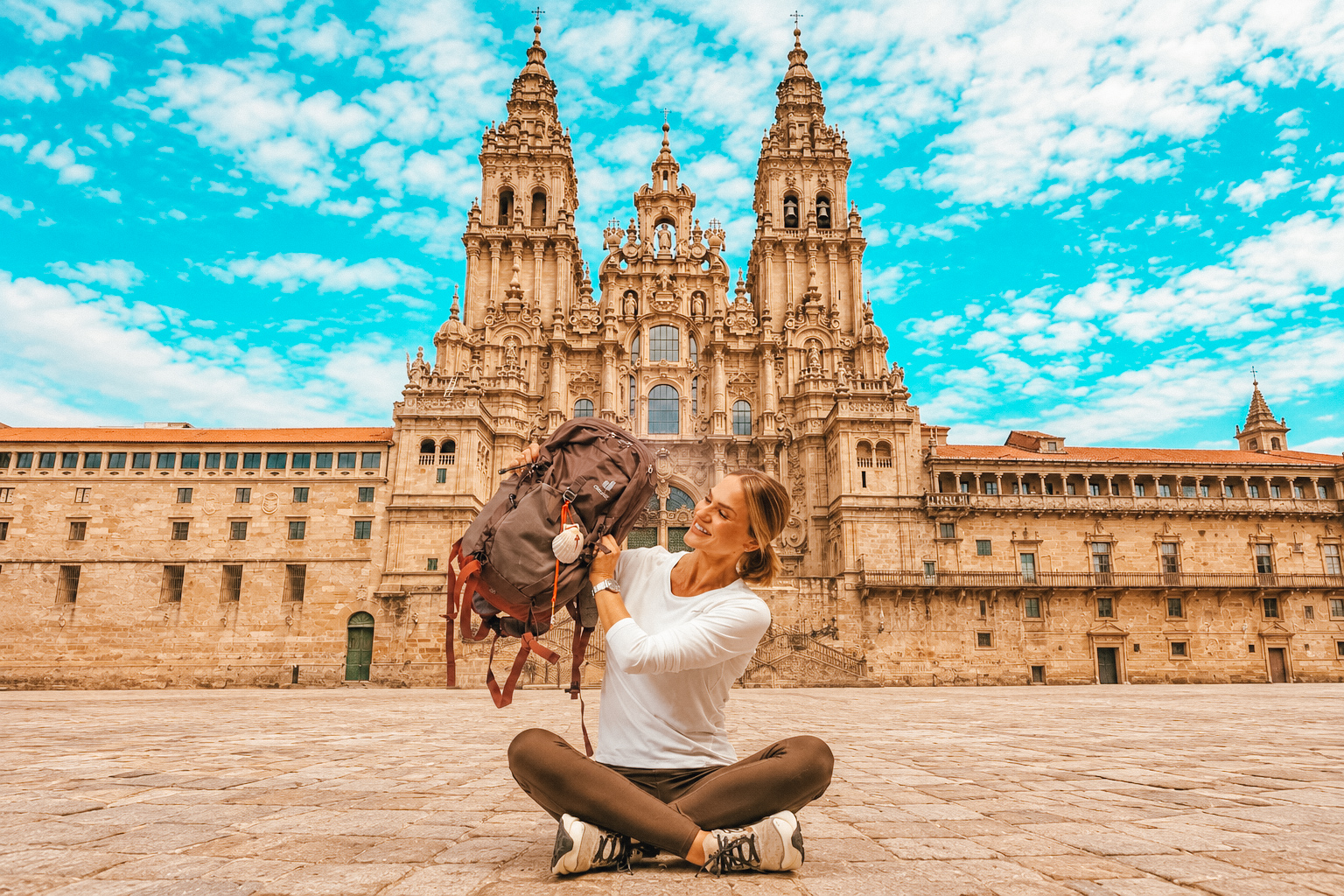 Emilim sentada no calçamento da Praça do Obradoiro, erguendo a mochila com a vieira, com a Catedral de Santiago de Compostela ao fundo