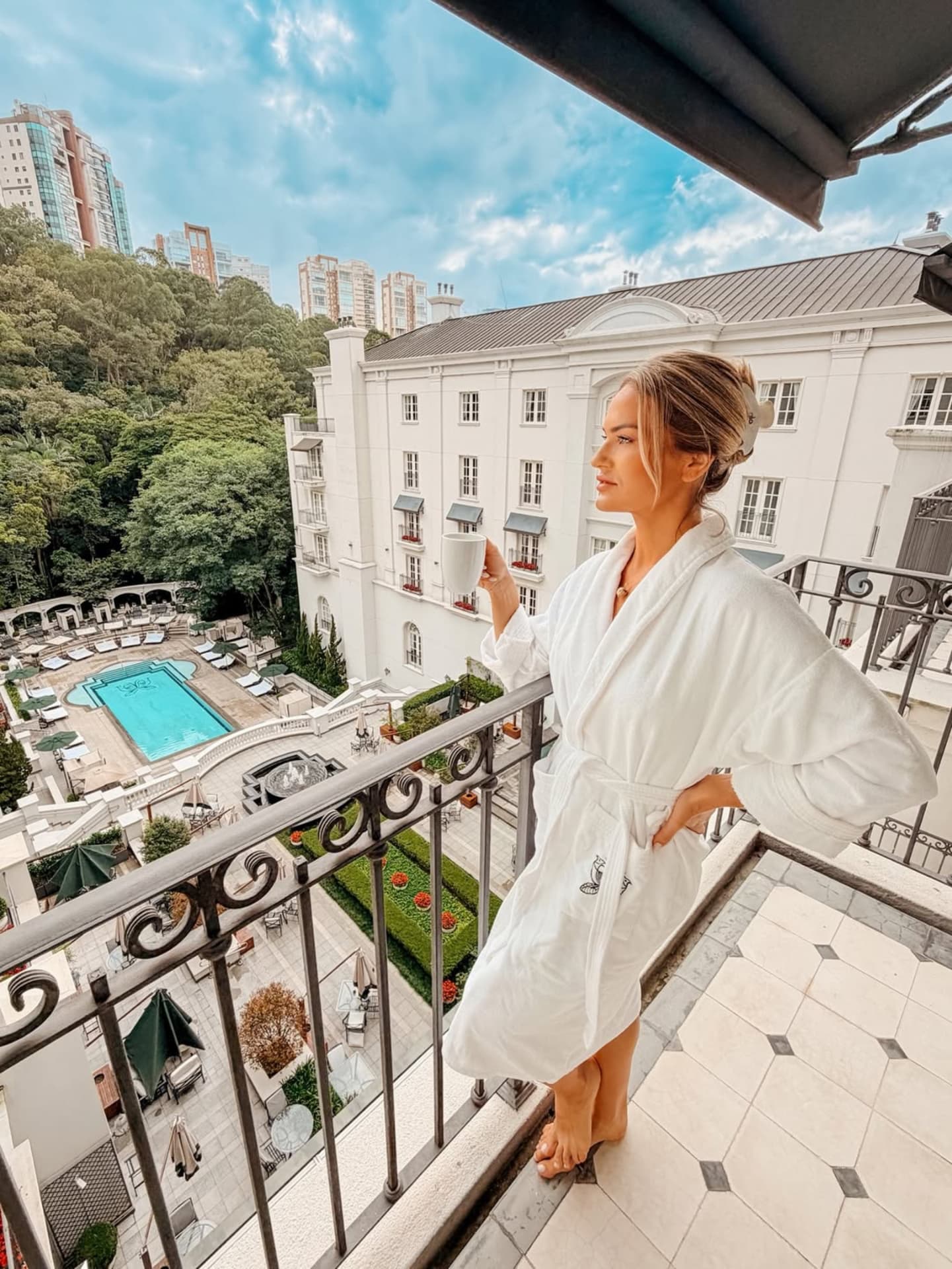 Emilim de roupão, de perfil, na varanda de um hotel, com a fachada branca, a piscina e os jardins ao fundo