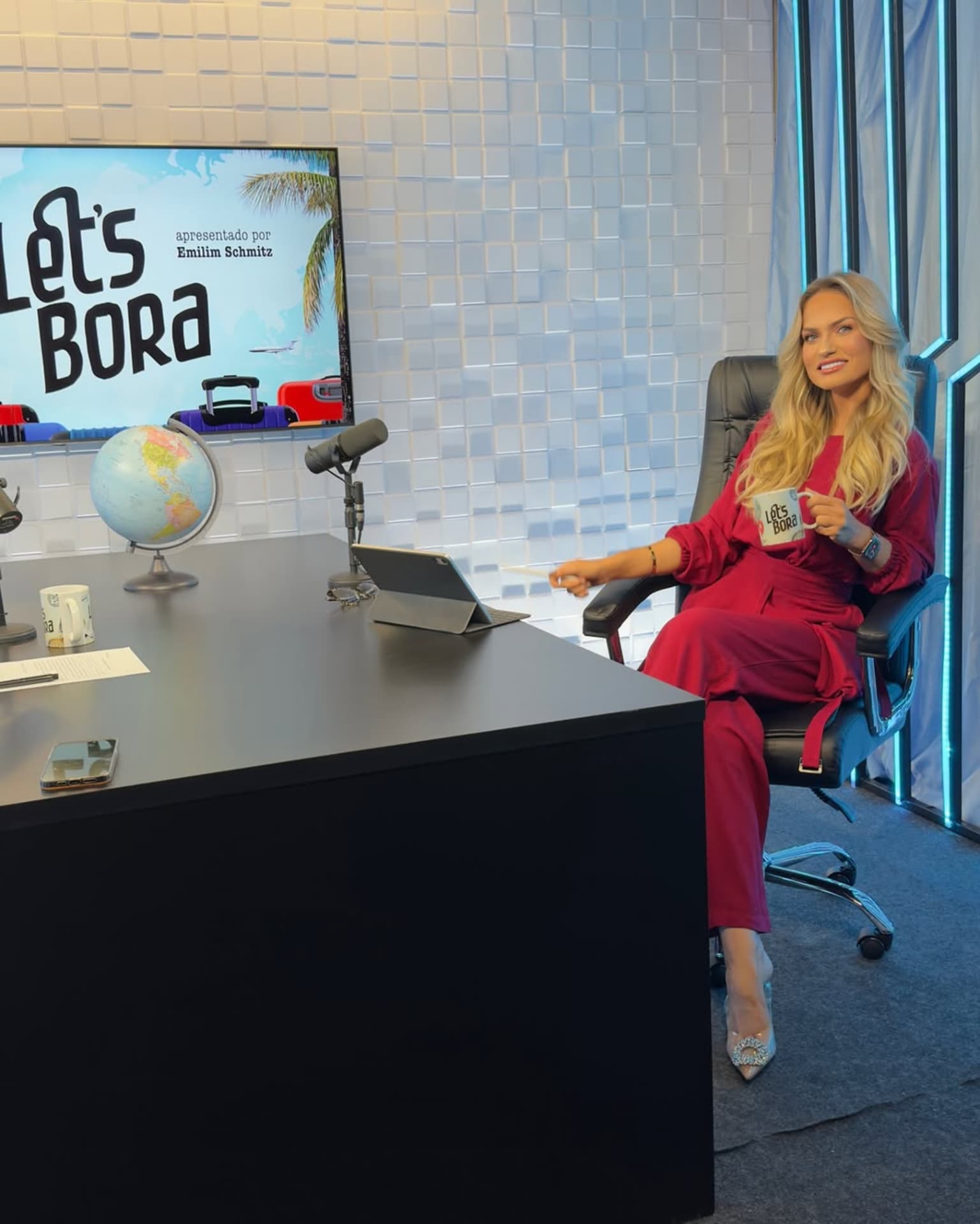 Emilim apresentando o programa Let's Bora, sentada à mesa do estúdio de TV com a caneca na mão